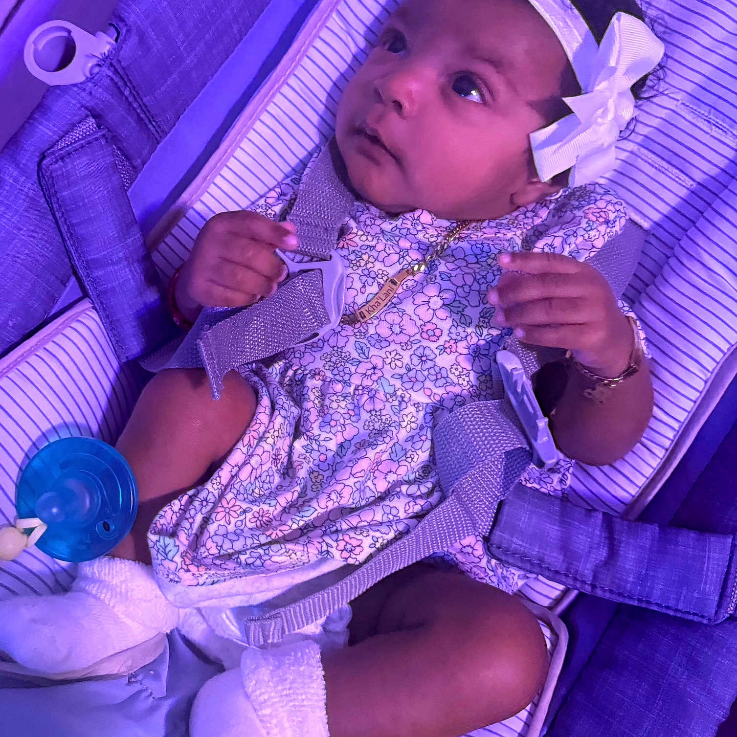 Khalani is registered to the contest to win money with this photo: baby, infant, child, headband, bow, floral_dress, white_socks, baby_carrier, straps, pacifier, purple_light, cute, gazing, curious, indoors, soft_lighting, closeup, portrait, comfort, resting
