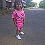 child, curly_hair, pink_outfit, shorts, sneakers, smile, outdoor, road, tree, building, grass, sidewalk, suburban, number_10, happy, person, casual_clothing, daylight, playful, young