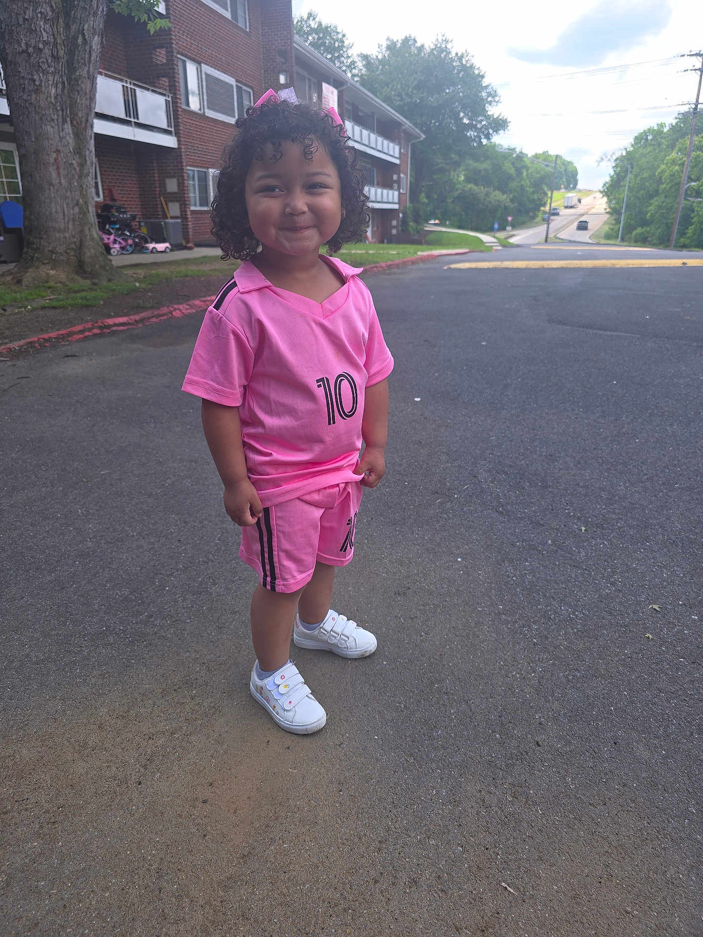 Sophia joined the competition — help win amazing prizes! child, curly_hair, pink_outfit, shorts, sneakers, smile, outdoor, road, tree, building, grass, sidewalk, suburban, number_10, happy, person, casual_clothing, daylight, playful, young