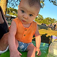 Maddox is registered to the contest to win money with this photo: baby, child, outdoor, sunlight, orange_clothing, blue_shorts, table, hand, face, person, nature, tree, grass, daylight, bottle, can, summer, curious, expression, park