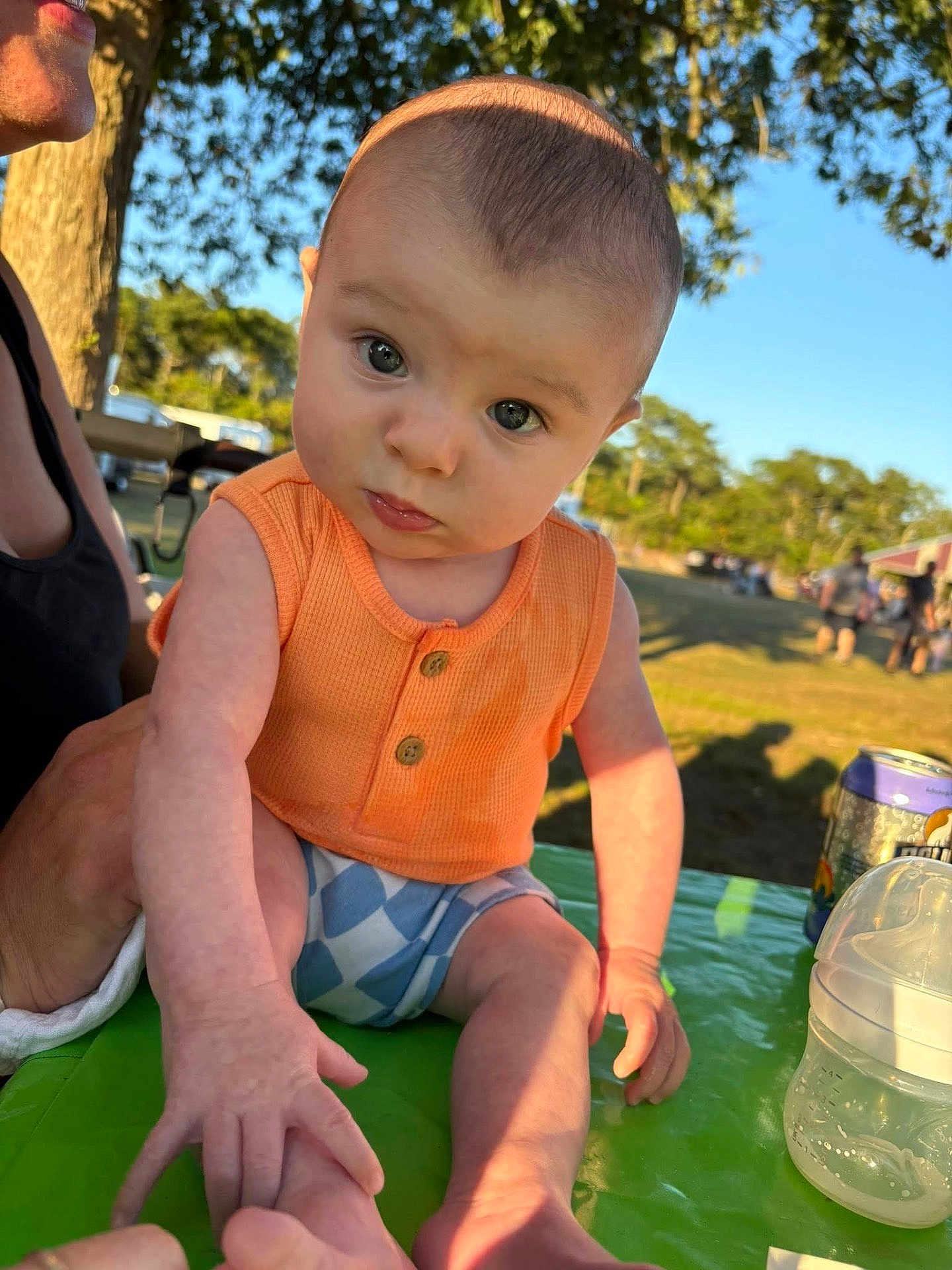 Maddox is registered to the contest to win money with this photo: baby, child, outdoor, sunlight, orange_clothing, blue_shorts, table, hand, face, person, nature, tree, grass, daylight, bottle, can, summer, curious, expression, park