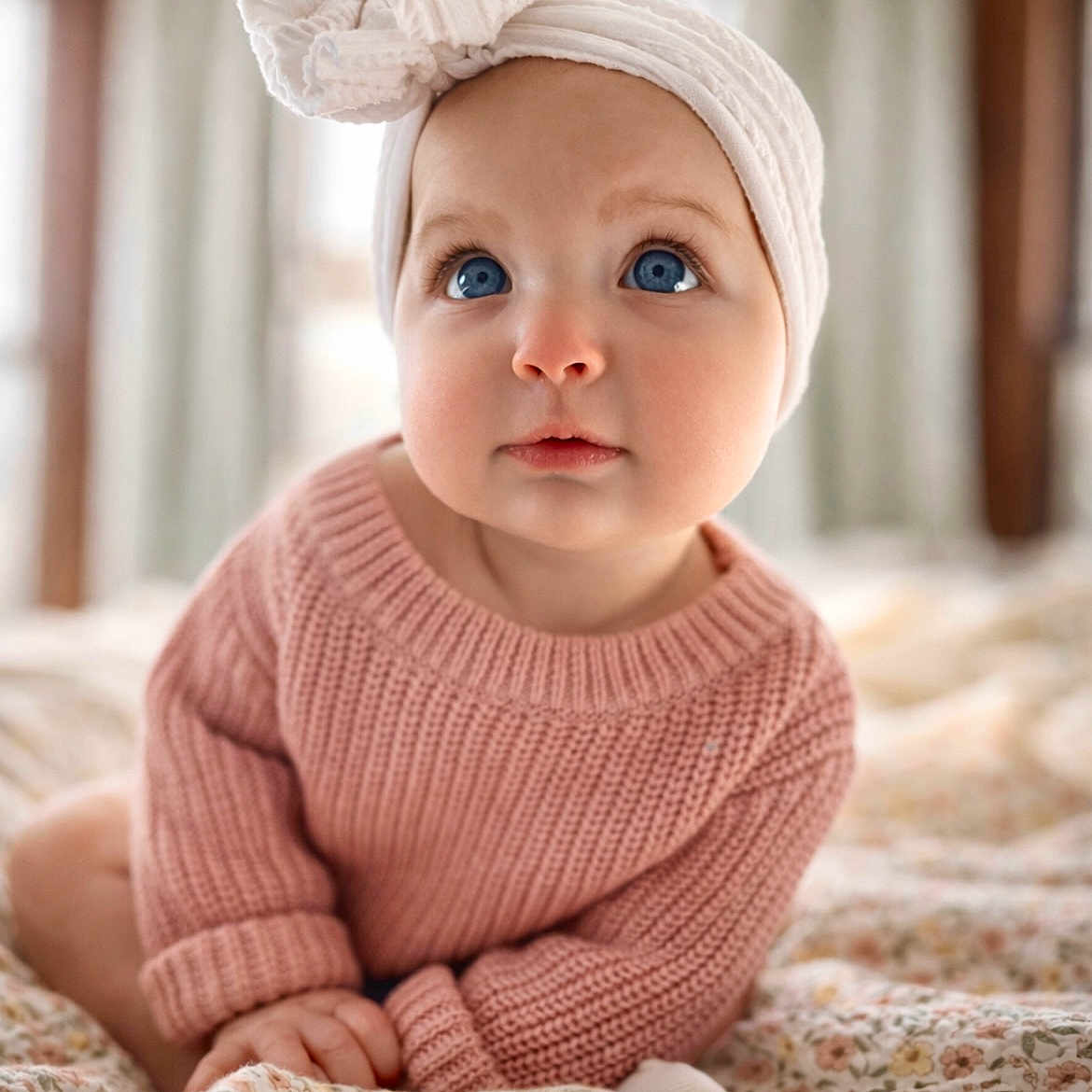 Juni is registered to the contest to win money with this photo: baby, bed, blanket, blue_eyes, child, cute, expression, face, floral_pattern, hands, headwrap, indoor, infant, knitwear, pink_sweater, portrait, sitting, soft_light, white_headband, window