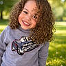 child, curly_hair, smiling, sweatshirt, car_graphic, outdoor, grass, sunlight, park, happy, portrait, face, person, casual_clothing, nature, daylight, young, cute, joy, leisure