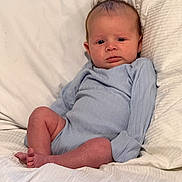 Cohan is registered to the contest to win money with this photo: baby, infant, blue_onesie, bed, blanket, pillow, person, skin, face, head, legs, feet, expression, indoors, sleepwear, newborn, cute, sitting, young_child, resting