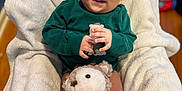Yeshua is registered to the contest to win money with this photo: baby, blanket, child, couch, cozy, cute, furniture, green_clothing, happy, indoor, infant, person, playful, plush_toy, portrait, red_scarf, smiling, soft_texture, teddy_bear, wooden_floor