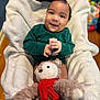 Yeshua is registered to the contest to win money with this photo: baby, smiling, green_clothing, blanket, teddy_bear, red_scarf, plush_toy, indoor, child, happy, person, couch, soft_texture, cute, portrait, infant, cozy, furniture, wooden_floor, playful