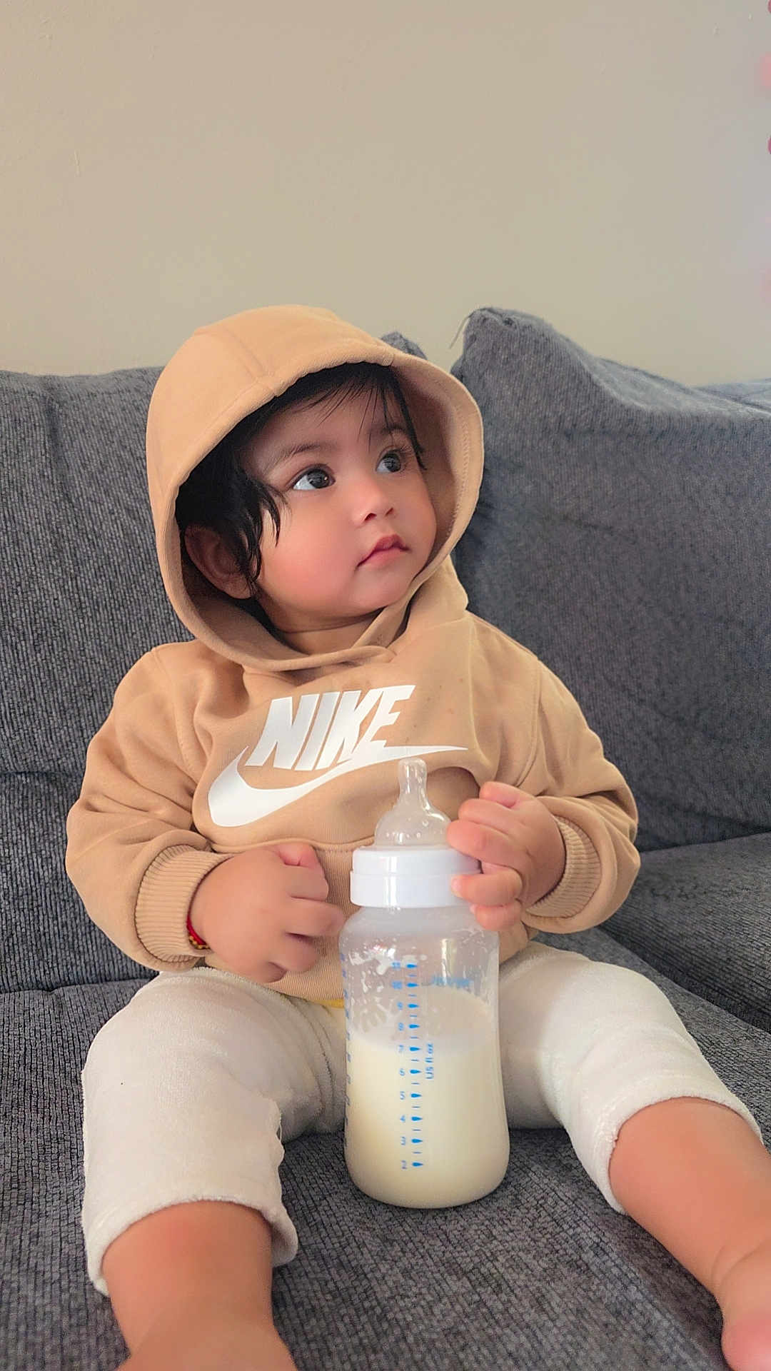 Sizda is registered to the contest to win money with this photo: toddler, child, hoodie, nike, milk_bottle, milk, sitting, couch, grey_couch, indoors, curious, clothing, baby, person, casual, cozy, soft_lighting, portrait, young_child, comfortable