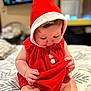 baby, bedspread, child, chubby_cheeks, cozy, curious, cute, face, hands, hood, indoors, infant, legs, long_eyelashes, pattern, pom_poms, red_clothing, sitting, skin, soft_fabric