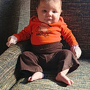 Presley is registered to the contest to win money with this photo: armchair, baby, barefoot, brown_pants, casual_clothing, child, cozy, cute, expression, face, furniture, indoor, infant, mittens, orange_shirt, person, portrait, seated, textured_fabric, young_child