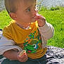toddler, child, outdoor, lake, grass, trees, sunlight, yellow_shirt, frog_design, snack, picnic_blanket, sitting, nature, daylight, cute, casual_clothing, barefoot, greenery, portrait, happy