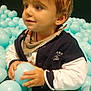 toddler, child, boy, ball_pool, plastic_balls, blue_balls, indoor, play_area, jacket, scarf, hands, face, hair, curious, seated, light_brown_hair, looking_away, soft_lighting, casual_clothing, playful