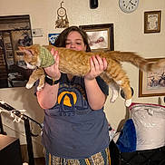 Han joined the competition — help win amazing prizes! person, cat, indoor, orange_cat, bandana, holding, portrait, clock, dreamcatcher, painting, wall_decor, watch, pajama_pants, home, furniture, relaxed, smile, long_cat, cozy, lights