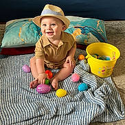 Kayden is registered to the contest to win money with this photo: baby, child, hat, straw_hat, smiling, sitting, blanket, easter_eggs, yellow_bucket, colorful, pillow, indoor, happy, toy, floor, cute, playful, young_child, infant, portrait
