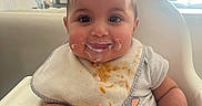 Delya participe au concours pour gagner de l'argent avec cette photo : baby, bib, high_chair, food, smile, chubby_cheeks, indoor, living_room, television, window, sunlight, clothing, cute, child, person, face, messy, happy, infant, feeding_time