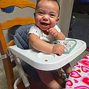 Luciano is registered to the contest to win money with this photo: baby, high_chair, smiling, indoor, table, tablecloth, heart_pattern, child, person, happy, clothing, seat, face, furniture, baby_bib, arm, hand, head, wooden_chair, floor