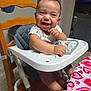 baby, high_chair, smiling, indoor, table, tablecloth, heart_pattern, child, person, happy, clothing, seat, face, furniture, baby_bib, arm, hand, head, wooden_chair, floor