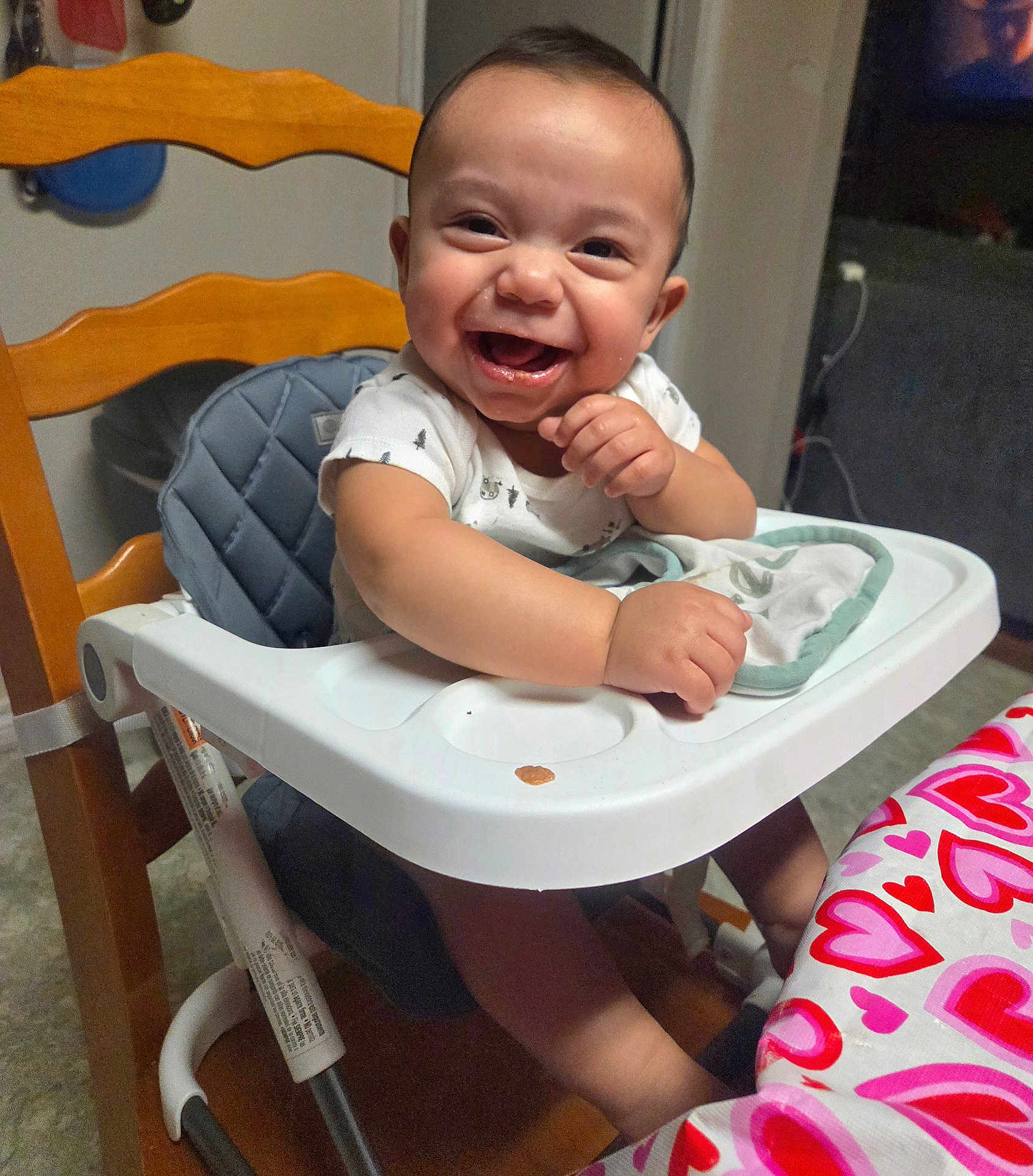 Luciano is registered to the contest to win money with this photo: baby, high_chair, smiling, indoor, table, tablecloth, heart_pattern, child, person, happy, clothing, seat, face, furniture, baby_bib, arm, hand, head, wooden_chair, floor