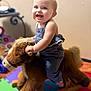 baby, child, smiling, rocking_horse, plush_toy, indoor, playroom, colorful, happy, cute, person, toy, floor_mat, blue_clothing, young_child, face, hands, seat, wooden_rocker, joy