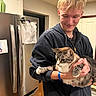 person, cat, tabby_cat, indoor, kitchen, refrigerator, hoodie, smile, blond_hair, facial_hair, holding, stretching_paw, appliance, paper, pin, cabinet, door, countertop, wood_floor, casual_clothing