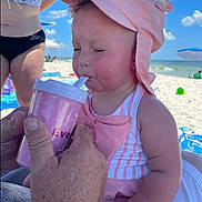 Everleigh is registered to the contest to win money with this photo: baby, child, beach, sun_hat, pink, cup, straw, striped_swimsuit, person, adult_hand, sky, cloud, sand, umbrella, vacation, summer, outdoor, water, relaxation, family