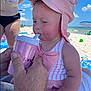 baby, child, beach, sun_hat, pink, cup, straw, striped_swimsuit, person, adult_hand, sky, cloud, sand, umbrella, vacation, summer, outdoor, water, relaxation, family