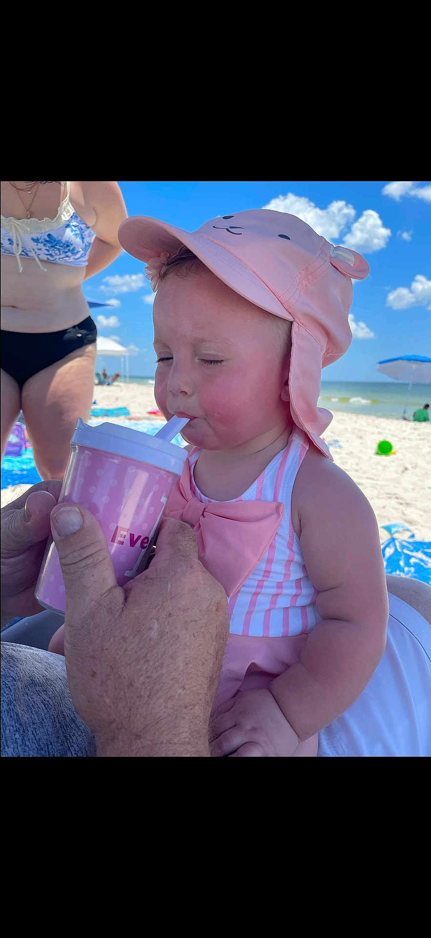 Everleigh is registered to the contest to win money with this photo: baby, child, beach, sun_hat, pink, cup, straw, striped_swimsuit, person, adult_hand, sky, cloud, sand, umbrella, vacation, summer, outdoor, water, relaxation, family