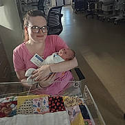 Mazikeen is registered to the contest to win money with this photo: baby, blanket, chair, cradle, glasses, holding, hospital, hospital_room, indoor, infant, medical_equipment, mother, newborn, nursery, patchwork_quilt, person, pink_shirt, smile, visitor_badge, woman