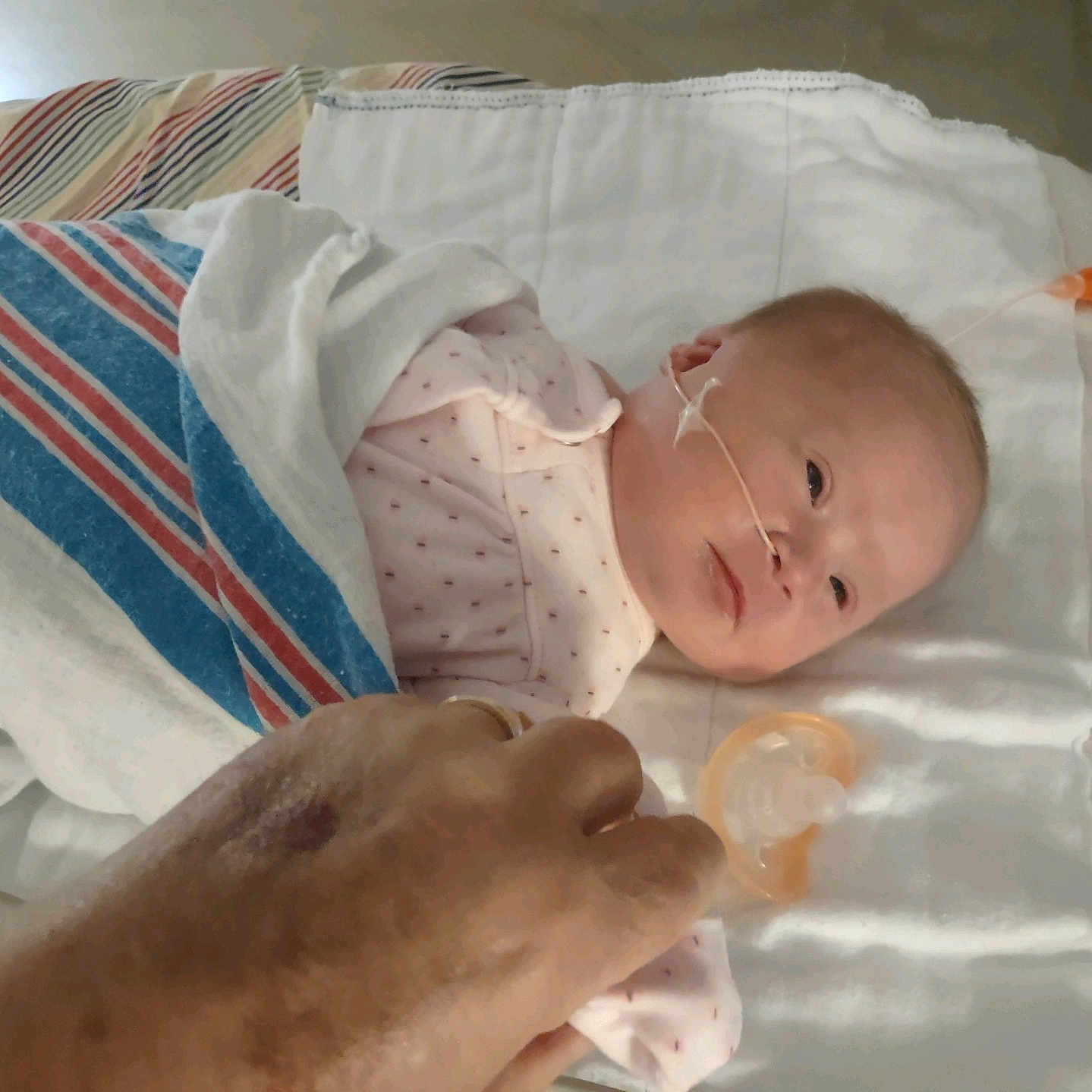 Mazikeen is registered to the contest to win money with this photo: baby, hospital, blanket, pacifier, nasal_tube, hand, medical, bed, infant, person, healthcare, newborn, care, resting, white_sheet, striped_blanket, pink_clothing, patient, treatment, closeup