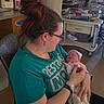 woman, newborn, baby, hospital, nursery, cradle, blanket, glasses, hair_bun, earring, tshirt, chair, medical_equipment, face, person, indoor, love, mother, holding, sleeping