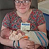 baby, infant, newborn, parent, woman, person, portrait, hospital, visitor_pass, blanket, swaddle, floral_clothing, glasses, smiling, sitting, chair, indoor, medical_bracelet, family, new_parent