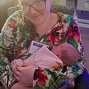 Mazikeen joined the competition — help win amazing prizes! baby, infant, adult, woman, person, holding, cradling, portrait, indoor, seated, glasses, smiling, floral_pattern, colorful_clothing, patterned_pants, blanket, visitor_badge, hospital, chair, family