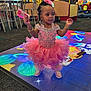 child, girl, pink_dress, tutu, hair_bow, indoor_play_area, toy, colorful_floor, led_floor, smiling, standing, playground, carpet, shoes, bracelet, happy, party_dress, beads, furniture, background