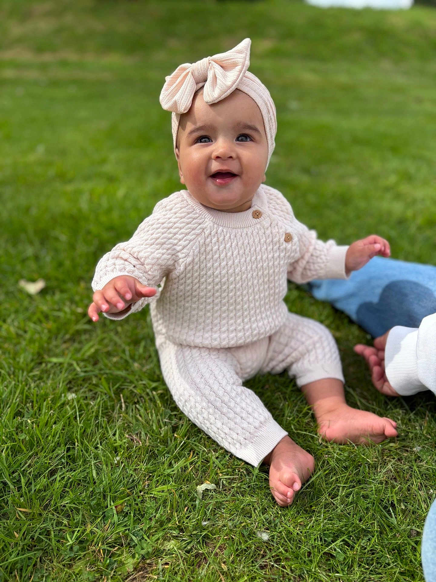 Billie joined the competition — help win amazing prizes! baby, child, grass, outdoor, knitted_clothing, headband, bow, smiling, sitting, person, barefoot, cute, happy, infant, nature, daylight, casual, young_child, greenery, supporting_hand