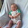 Ozlo is registered to the contest to win money with this photo: baby, smiling, infant, white_overalls, green_shirt, blanket, soft_texture, sitting, happy, cute, child, portrait, indoors, natural_light, skin, hands, feet, face, person, cozy