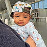 arm, baby, blue_outfit, cap, child, closeup, clothing, curious_expression, cute, dinosaur_print, face, headwear, holding, indoor, infant, modern_building, person, portrait, sailboat_print, window