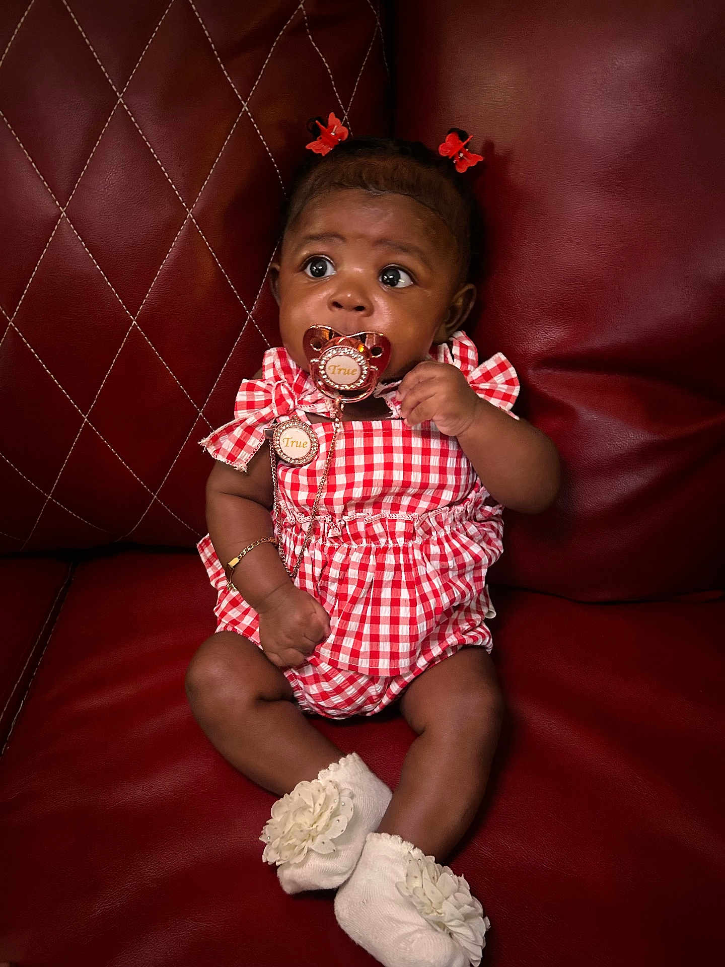 True is registered to the contest to win money with this photo: baby, child, pacifier, red_leather_couch, checkered_outfit, socks, flower_decoration, hair_clips, infant, cute, sitting, indoor, portrait, adorable, young_child, skin, face, expression, clothing, furniture