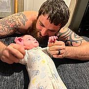 Jake is registered to the contest to win money with this photo: baby, man, tattoo, beard, pajamas, indoor, bedroom, holding_hands, infant, cute, love, family, smile, person, face, carpet, door, hand, interaction, lying_down