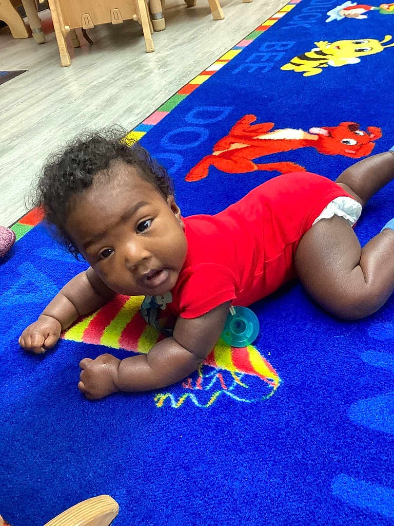 William joined the competition — help win amazing prizes! baby, child, infant, red_onesie, blue_carpet, cartoon_characters, pacifier, floor, wooden_chair, curly_hair, indoors, playroom, toddler, colorful, carpet_pattern, crawling, cute, young_child, expression, furniture