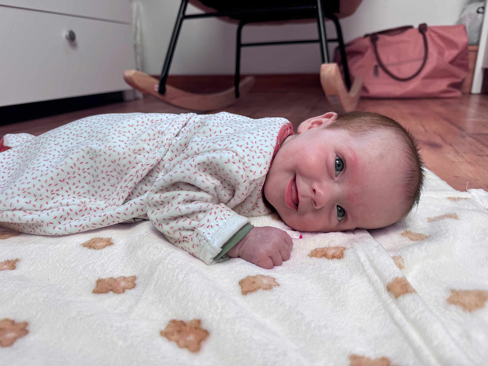 Lya participe au concours pour gagner de l'argent avec cette photo : baby, infant, smiling, blanket, teddy_bear_pattern, floor, wooden_floor, rocking_chair, pink_bag, face, hand, clothing, long_sleeve, indoor, cute, child, portrait, resting, tongue_out, lying_down