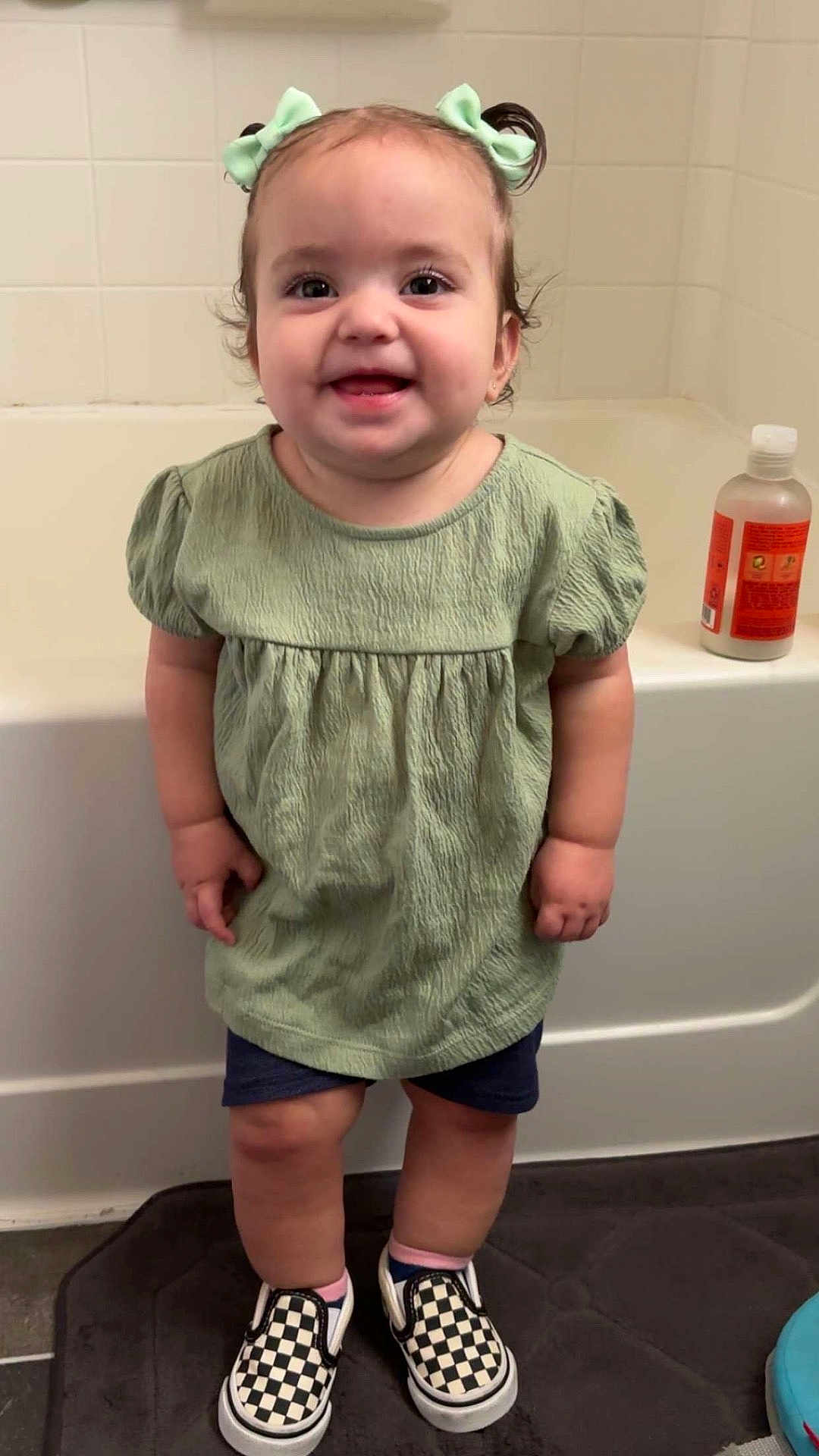 Raelynn is registered to the contest to win money with this photo: toddler, child, smile, green_dress, hair_bows, checkered_shoes, bathroom, bathtub, floor_mat, shorts, standing, happy, indoor, person, cute, small_hands, wall_tiles, skin, socks, eyebrows