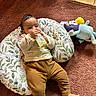 baby, bottle, brown_hair, carpet, child, clothing, cozy, cushion, dragon_toy, face, feeding, floor, home, indoor, infant, lying_down, plush_toy, portrait, sitting, sunlight