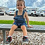 toddler, child, bench, denim_overalls, sneakers, nike, curly_hair, outdoor, parking_lot, gravel, grass, cloudy_sky, cars, metal_bench, casual_clothing, person, summer, daylight, portrait, cute