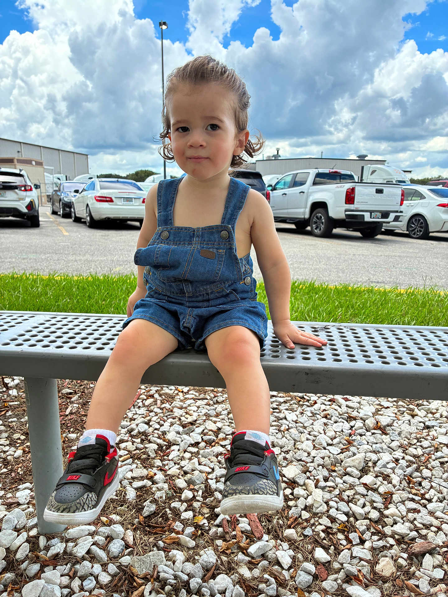Echeniel is registered to the contest to win money with this photo: toddler, child, bench, denim_overalls, sneakers, nike, curly_hair, outdoor, parking_lot, gravel, grass, cloudy_sky, cars, metal_bench, casual_clothing, person, summer, daylight, portrait, cute
