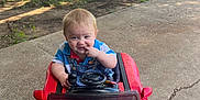 Noah is registered to the contest to win money with this photo: toddler, child, toy_car, red_car, outdoor, sidewalk, grass, building, playful, blonde_hair, concrete, daylight, person, casual_clothing, smiling, fun, summer, nature, cute, small_child