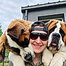 person, dogs, puppies, saint_bernard, pet, animal, selfie, smiling, sunglasses, baseball_cap, outdoor, house, sky, cloudy, fence, fur, white_and_brown, large_dogs, companionship, portrait
