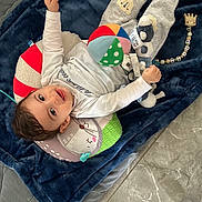 Rayan a rejoint le concours — aidez-le/la à gagner de superbes lots ! baby, child, toy, blanket, plush_toy, ball, floor, slipper, smiling, clothing, pillow, indoor, cute, infant, happy, socks, person, head, face, lying_down