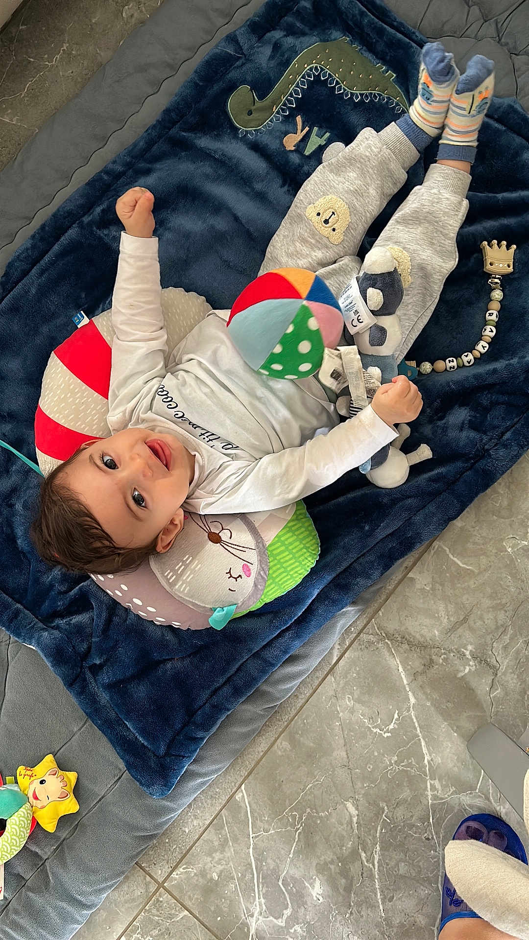 Rayan a rejoint le concours — aidez-le/la à gagner de superbes lots ! baby, child, toy, blanket, plush_toy, ball, floor, slipper, smiling, clothing, pillow, indoor, cute, infant, happy, socks, person, head, face, lying_down