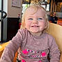 blond_hair, blue_eyes, casual, child, clothing, cute, face, hand, happy, high_chair, indoor, person, pink_shirt, portrait, restaurant, seat_belt, smiling, table, tassels, toddler