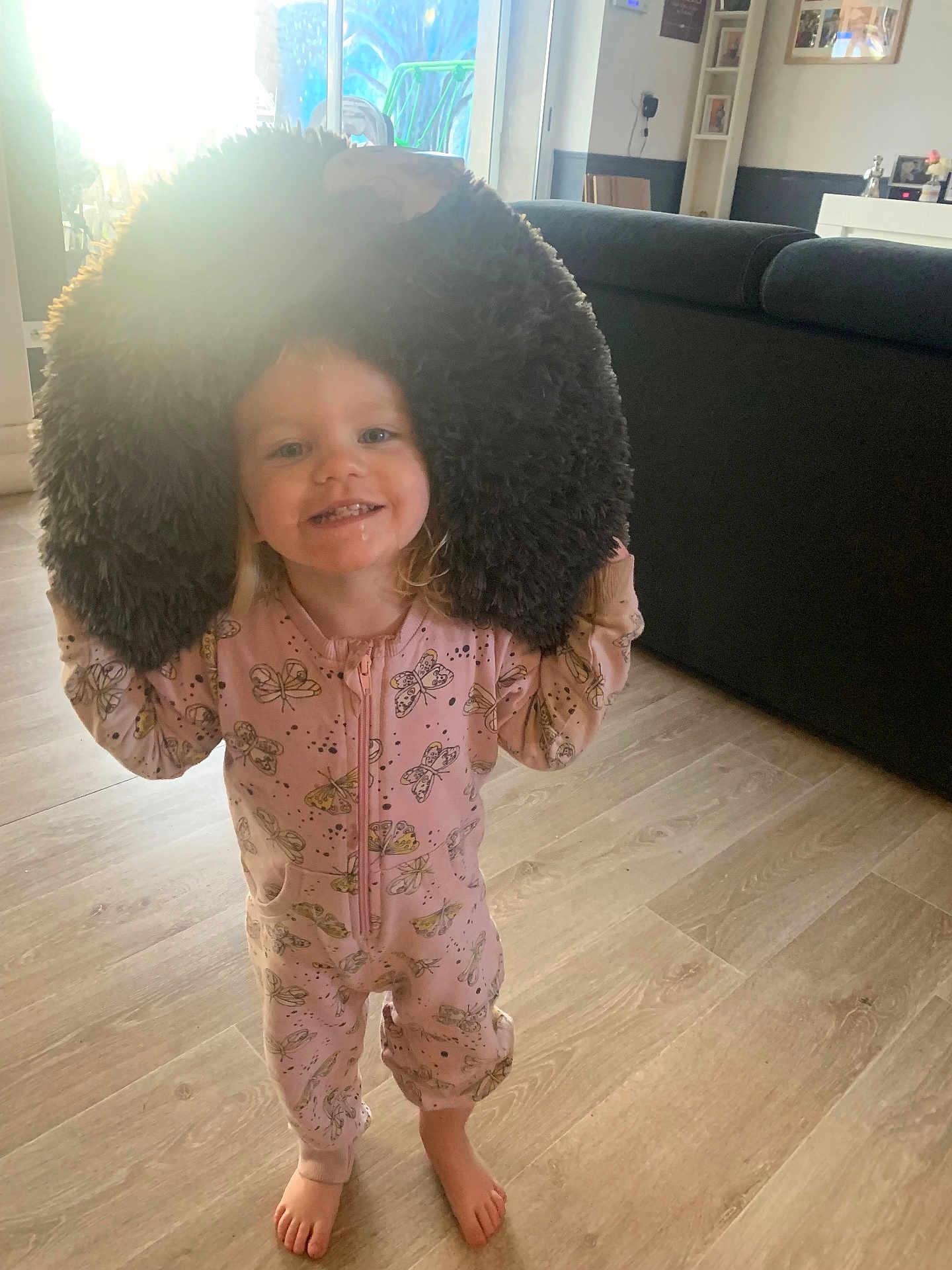 Ella participe au concours pour gagner de l'argent avec cette photo : barefoot, butterfly_pattern, child, couch, floor, fluffy, happy, home, indoor, living_room, onesie, pajamas, person, pillow, playful, smiling, sunlight, toddler, window, wooden_floor