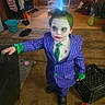 child, face_paint, joker_costume, purple_suit, green_tie, indoor, messy_hair, carpet, toy, bucket, broom, bag, milk_crate, child_costume, black_shoes, clutter, person, portrait, costume, young_child